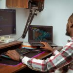 An African man coding on a desktop and laptop in a Nairobi office setting, showcasing modern technology.