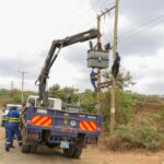 Utility workers in protective gear servicing power lines with a truck crane on a rural road.